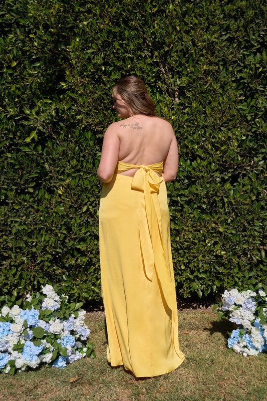 Back view of plus size yellow bridesmaid dress featuring open back, bow tie closure, long flowing ribbons and floor-length silhouette.