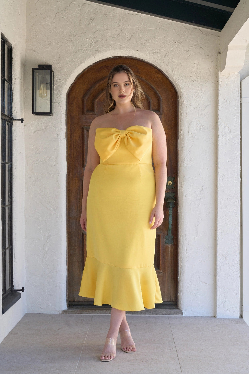 Plus size bright yellow strapless wedding guest dress with oversized bow neckline, fitted mermaid silhouette and flared tea-length hem.