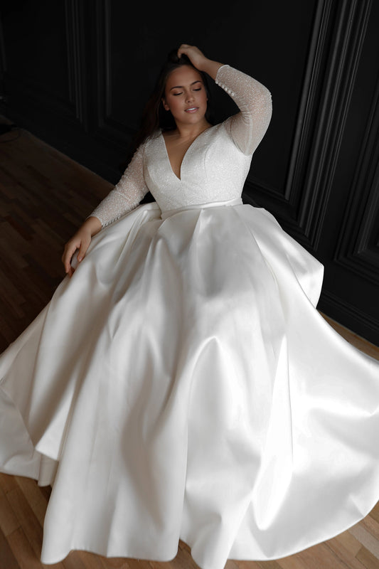 White plus size ball gown wedding dress with sparkling plunging V-neck bodice, sheer glitter long sleeves and voluminous satin-look skirt